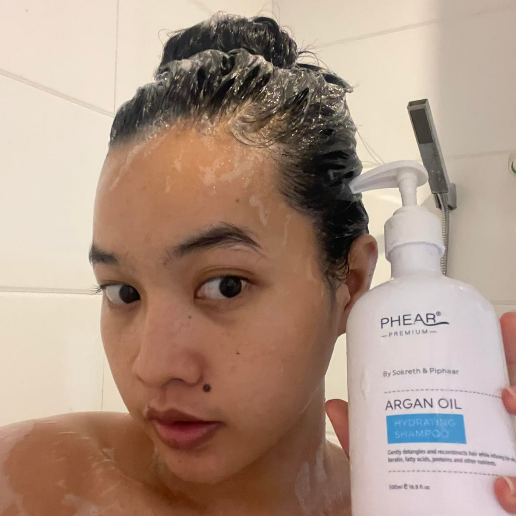 A beautiful woman with shampoo on head holding a bottle of PHEAR Argan Oil shampoo.